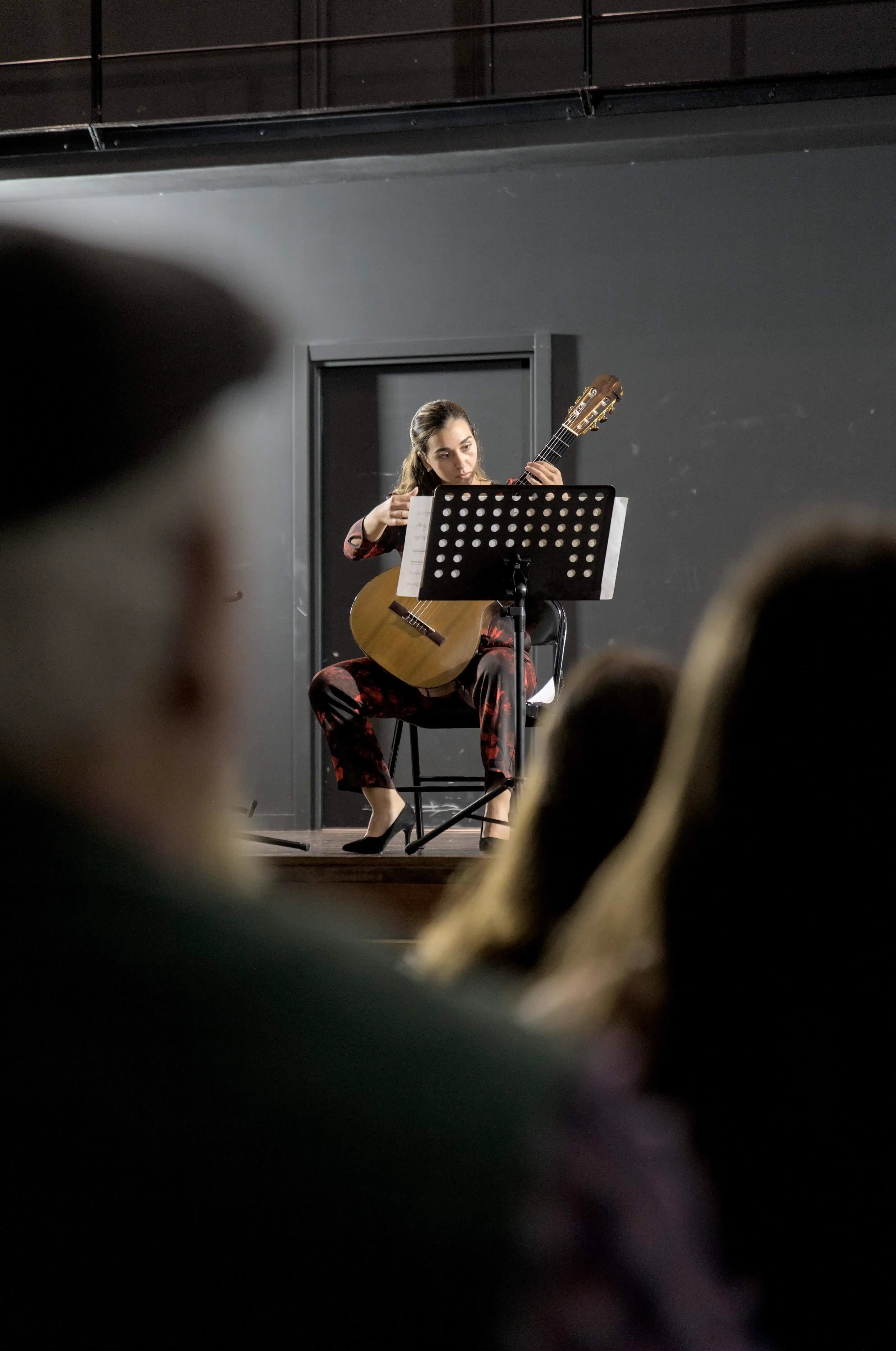 Uma mulher toca guitarra clássica enquanto olha a partitura. Em primeiro plano vê-se as pessoas da plateia.